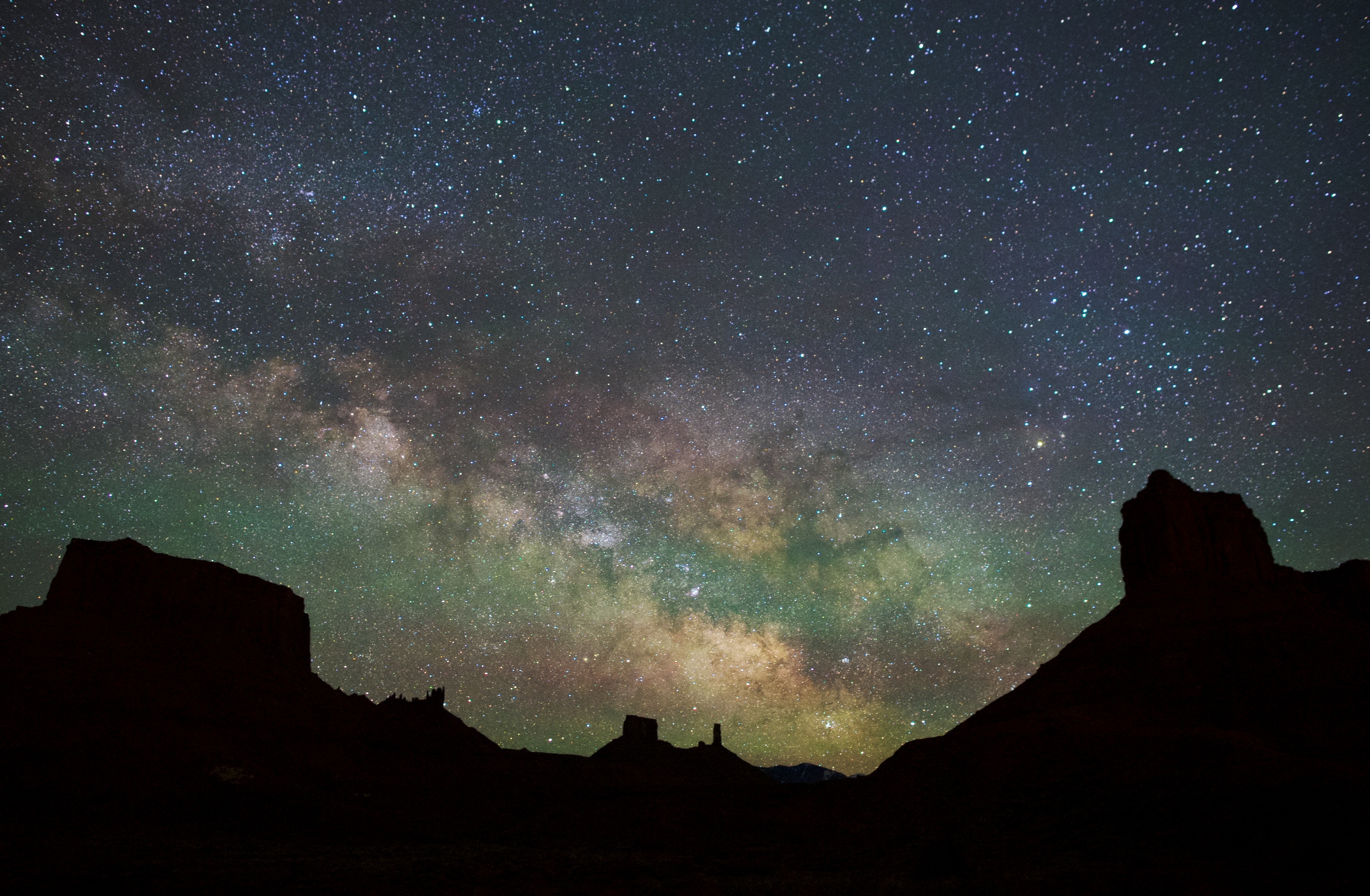 a dark horizon at night, with huge slihouettes of sandstone mesas. the bright Milky Way arches across the sky and bands green glow pass in front.