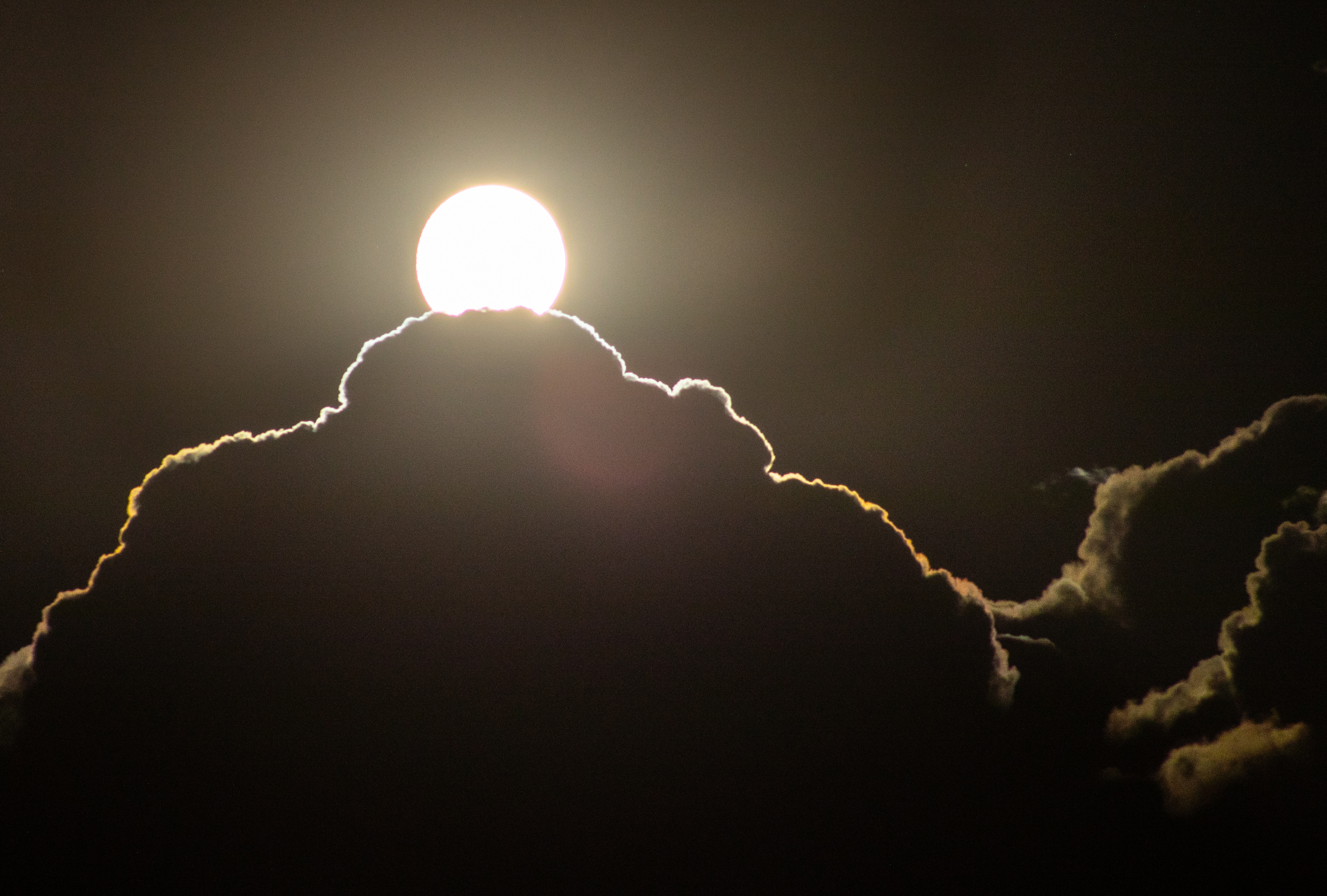 a bright full moon peeks out from a bank of clouds lined in silver moonlight