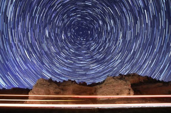 long-exposure photo of the sky and a highway and cliffs shows the stars as circular glowing trails and passing cars as streaks of light